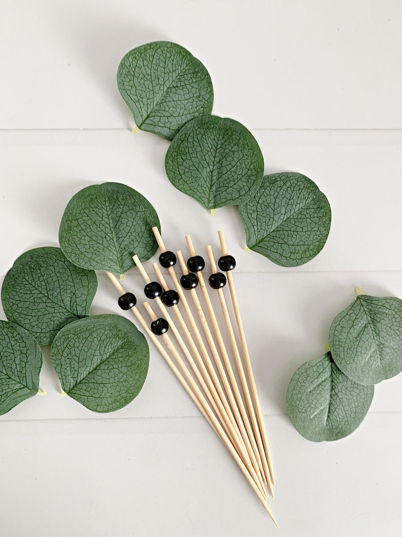 May include: A collection of bamboo skewers topped with black beads, arranged with green eucalyptus leaves. The skewers are natural wood color, and the leaves are a deep green. The arrangement is on a white surface.
