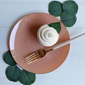 May include: A frosted cupcake with a green leaf garnish sits on a pink plate with a gold rim. A gold fork and a white-handled spoon are placed on the plate. Green leaves are arranged around the plate on a white wooden surface.