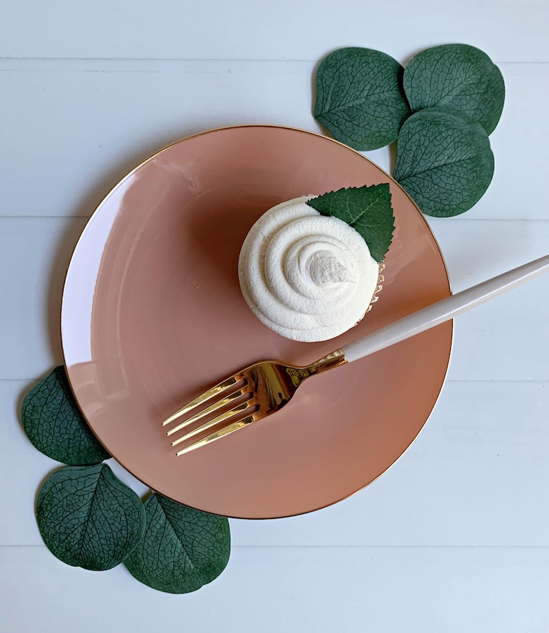 May include: A frosted cupcake with a green leaf garnish sits on a pink plate with a gold rim. A gold fork and a white-handled spoon are placed on the plate. Green leaves are arranged around the plate on a white wooden surface.