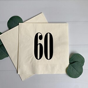 May include: Two square cream-coloured paper napkins featuring the number 60 printed in a bold black serif font centred on the front. The napkins have a textured border along the edges and are accented by dark green eucalyptus leaves.