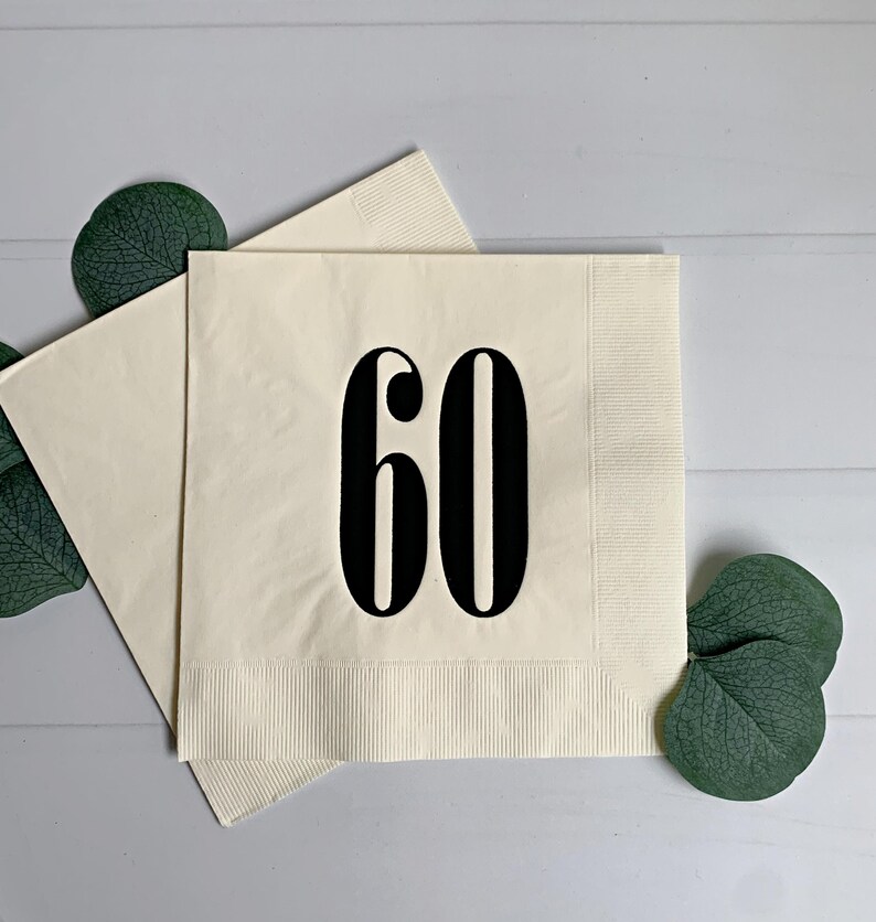 May include: Two square cream-coloured paper napkins featuring the number 60 printed in a bold black serif font centred on the front. The napkins have a textured border along the edges and are accented by dark green eucalyptus leaves.