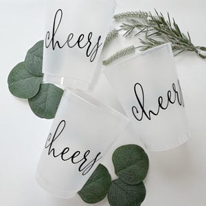 May include: Three frosted plastic cups with the word "cheers" in black script. The cups are surrounded by green eucalyptus leaves and a sprig of greenery. Perfect for a party or celebration.