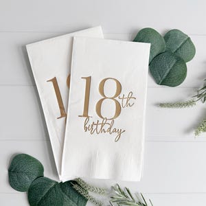 May include: Two white paper napkins with gold text that reads "18th birthday". The napkins are arranged on a white surface with green decorative leaves and sprigs.
