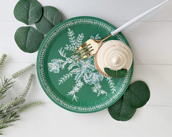 May include: A green paper plate with a white floral design. A cupcake with white frosting is on the plate, and a gold fork is next to it.