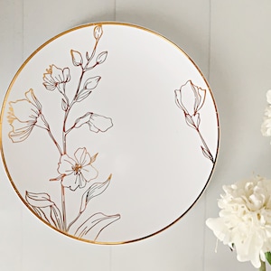 May include: A round white ceramic plate featuring a delicate gold-colored line drawing of a floral stem with leaves and blossoms. The plate has a thin gold-colored rim, creating a clean and polished appearance for this dining piece.