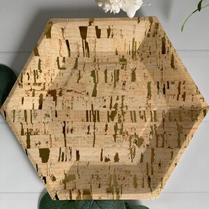 May include: A hexagonal paper plate with a cork-like design and gold accents. The plate has a natural, textured appearance with a light brown base and irregular gold streaks. Decorative white flowers and green leaves are in the background.
