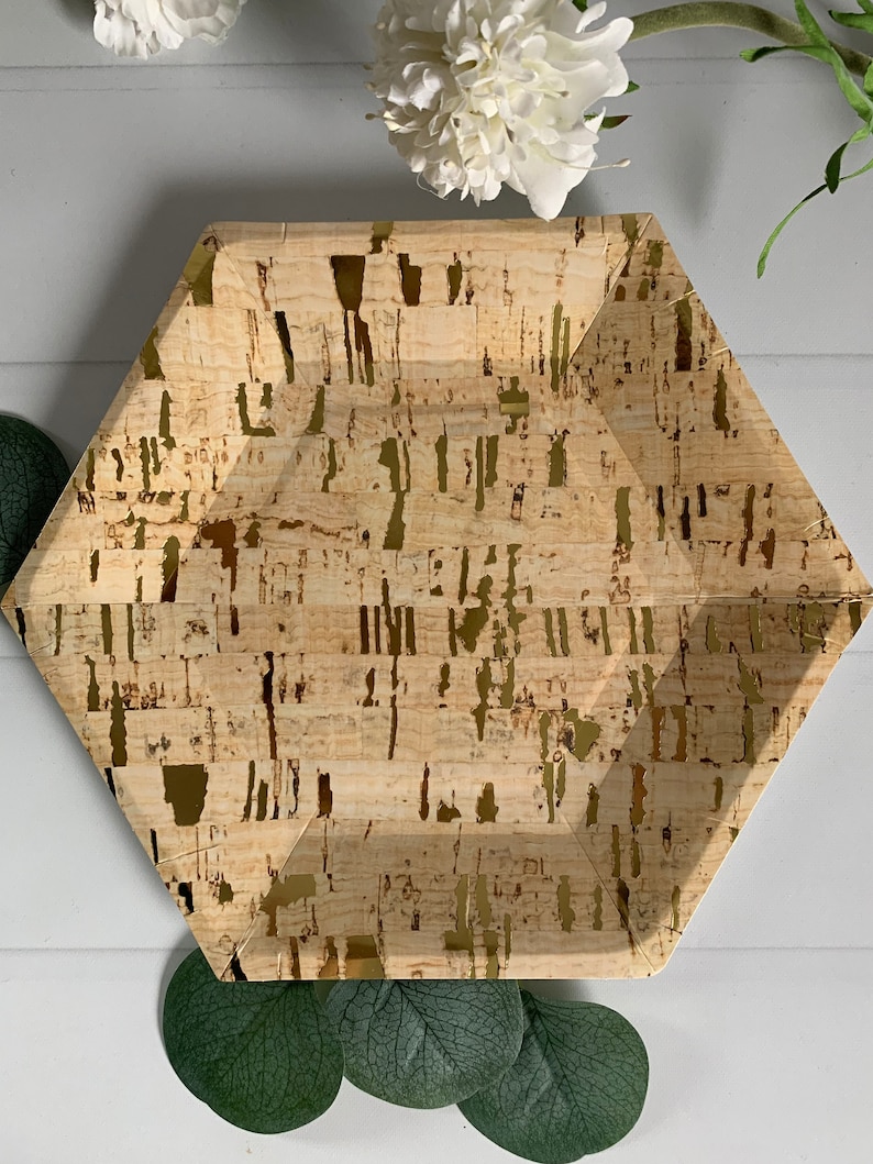 May include: A hexagonal paper plate with a cork-like design and gold accents. The plate has a natural, textured appearance with a light brown base and irregular gold streaks. Decorative white flowers and green leaves are in the background.