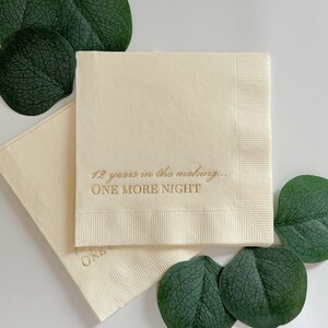 May include: Two square cream-colored paper napkins featuring gold foil text that reads 12 years in the making... ONE MORE NIGHT. The napkins are layered and surrounded by several dark green eucalyptus leaves on a plain white surface.