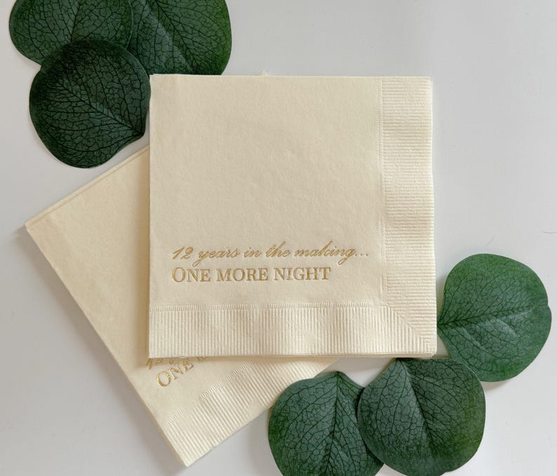May include: Two square cream-colored paper napkins featuring gold foil text that reads 12 years in the making... ONE MORE NIGHT. The napkins are layered and surrounded by several dark green eucalyptus leaves on a plain white surface.