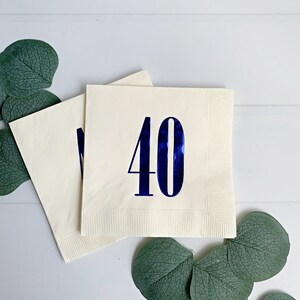 May include: Two white napkins with blue foil numbers 40 printed on them. The napkins are folded and placed on a white surface with green eucalyptus leaves.