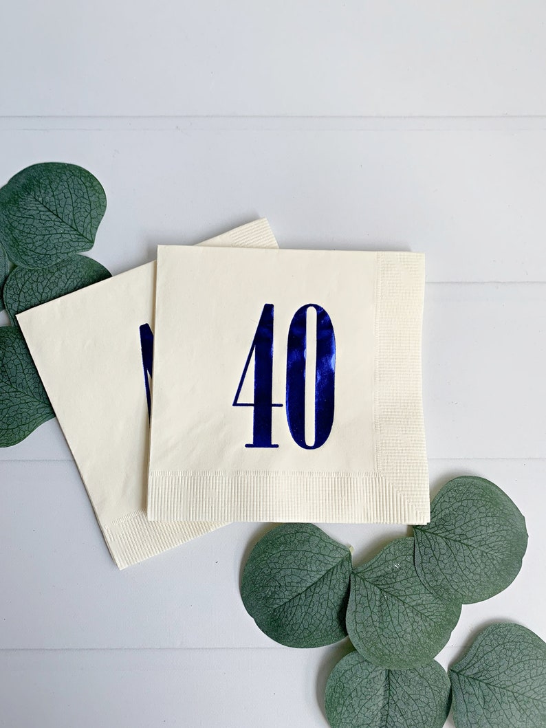 May include: Two white napkins with blue foil numbers 40 printed on them. The napkins are folded and placed on a white surface with green eucalyptus leaves.