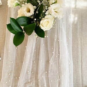 Pu&ograve; includere: Tessuto di tulle bianco con piccoli abbellimenti di perle bianche. Il tessuto &egrave; drappeggiato su una superficie marrone chiaro e un bouquet di fiori bianchi &egrave; sopra il tessuto.