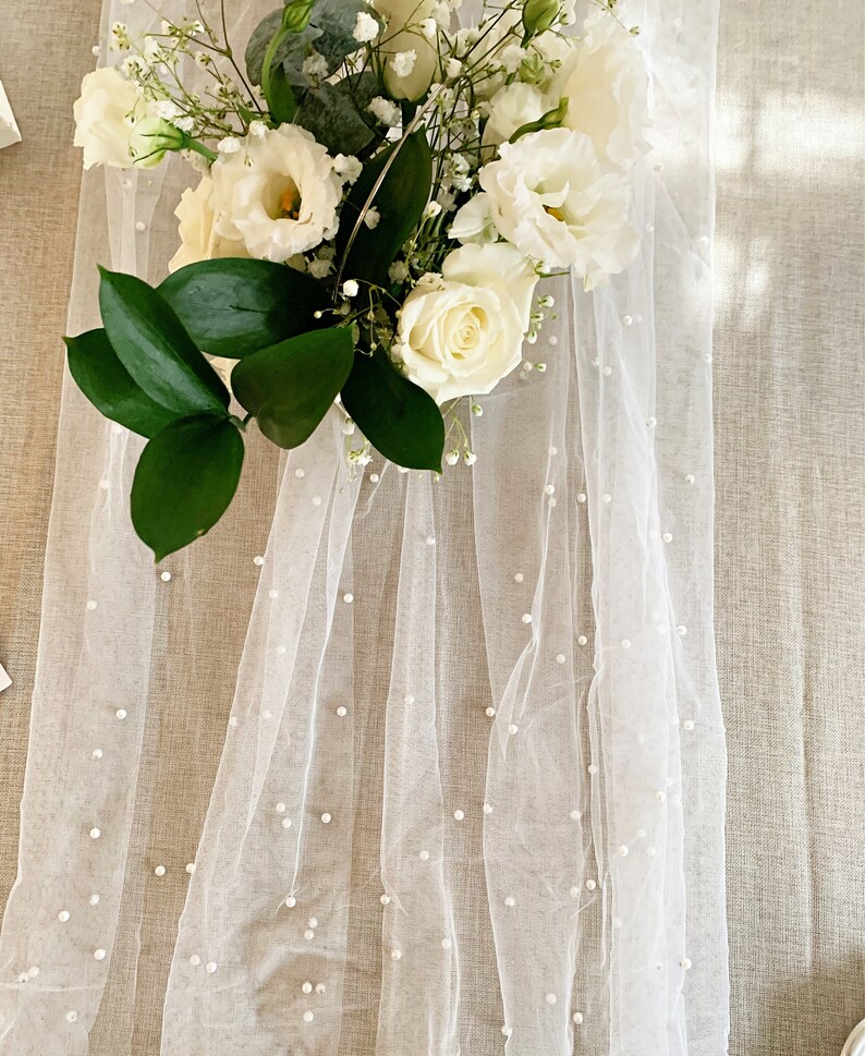 Pu&ograve; includere: Tessuto di tulle bianco con piccoli abbellimenti di perle bianche. Il tessuto &egrave; drappeggiato su una superficie marrone chiaro e un bouquet di fiori bianchi &egrave; sopra il tessuto.