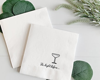 May include: Two white paper napkins with a black cocktail glass and the text "The Night Before..." printed on one of the napkins. The napkins are on a white surface with green eucalyptus leaves.