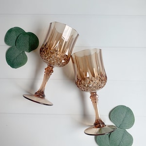 Op de afbeelding: Twee amberkleurige wijnglazen met diamantgeslepen details. De glazen hebben lange stelen en ronde voetjes. Groene bladeren zijn rond de glazen gerangschikt op een wit houten oppervlak. De glazen staan schuin naar elkaar toe.