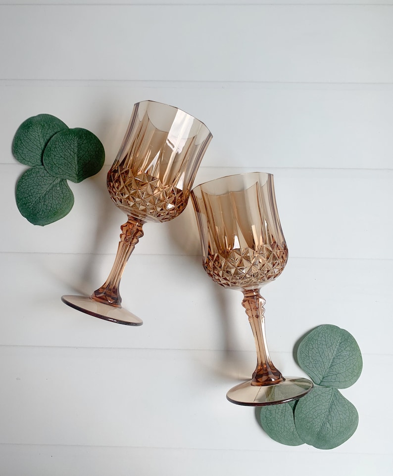 Op de afbeelding: Twee amberkleurige wijnglazen met diamantgeslepen details. De glazen hebben lange stelen en ronde voetjes. Groene bladeren zijn rond de glazen gerangschikt op een wit houten oppervlak. De glazen staan schuin naar elkaar toe.