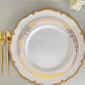 May include: A white dinner plate with a scalloped edge and a gold rim, featuring a delicate gold floral pattern and a soft pink band. The plate is accompanied by a gold-toned fork, knife, and spoon arranged neatly on the sides.