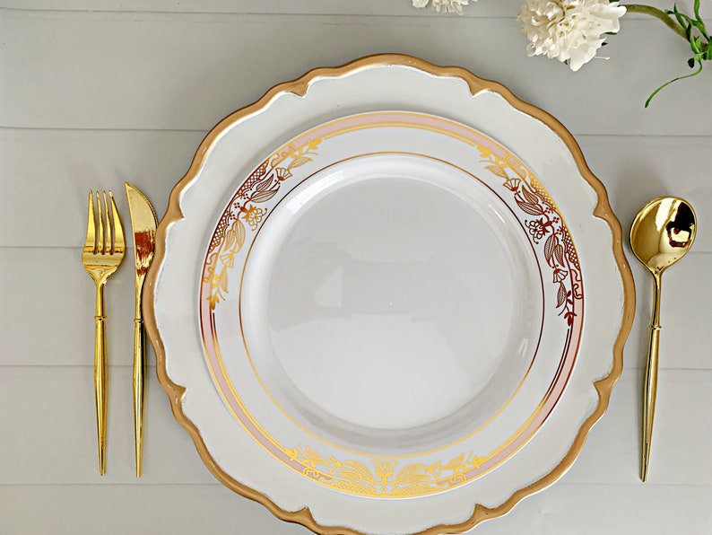 May include: A white dinner plate with a scalloped edge and a gold rim, featuring a delicate gold floral pattern and a soft pink band. The plate is accompanied by a gold-toned fork, knife, and spoon arranged neatly on the sides.