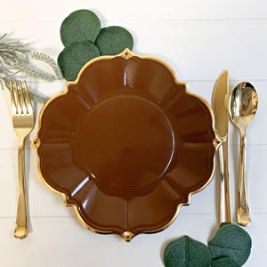 May include: A brown scalloped plate with a gold rim, accompanied by gold cutlery: a fork, knife, and spoon. Green leaves and sprigs of greenery are arranged around the plate setting on a white wooden surface.