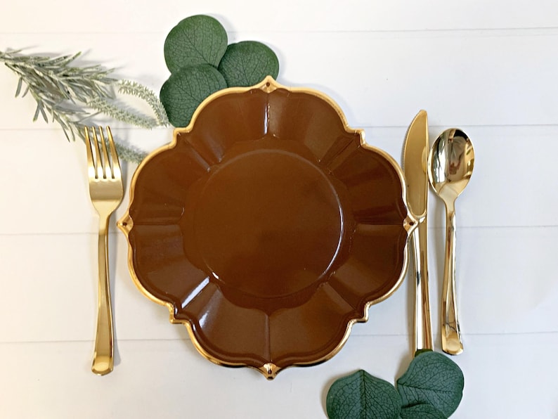 May include: A brown scalloped plate with a gold rim, accompanied by gold cutlery: a fork, knife, and spoon. Green leaves and sprigs of greenery are arranged around the plate setting on a white wooden surface.