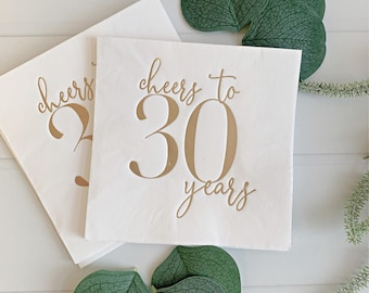 May include: Two square white paper napkins featuring the gold foil text "cheers to 30 years" printed in a mix of script and serif fonts. The napkins are arranged with several dark green eucalyptus leaves placed around the edges.