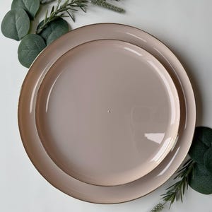 May include: Two blush-colored ceramic plates, one nested inside the other, with a gold rim. The plates are surrounded by green foliage. The plates are a soft, neutral color, suitable for a variety of table settings.