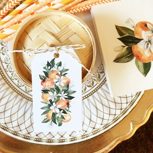 Op de afbeelding: Een decoratieve opstelling met een bamboe mand, oranje en wit gestreepte rietjes en een label met een aquarelontwerp van sinaasappels en bloemen. Op het label staat "dank u". De items staan op een goudkleurige plaat.