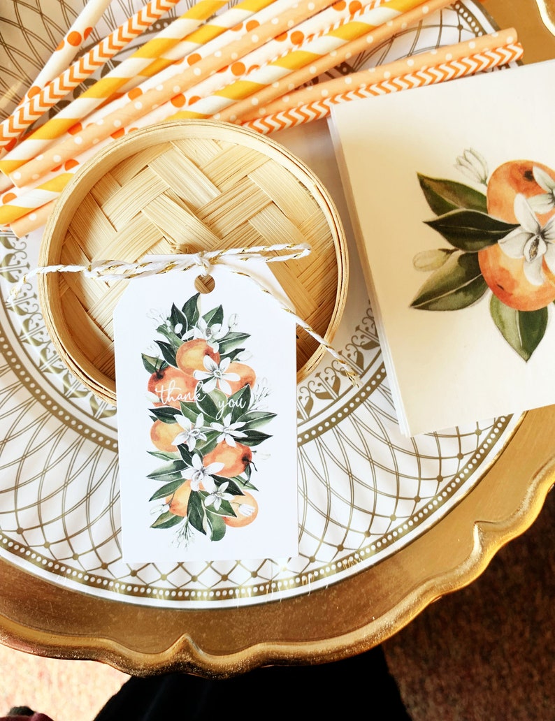 Op de afbeelding: Een decoratieve opstelling met een bamboe mand, oranje en wit gestreepte rietjes en een label met een aquarelontwerp van sinaasappels en bloemen. Op het label staat "dank u". De items staan op een goudkleurige plaat.
