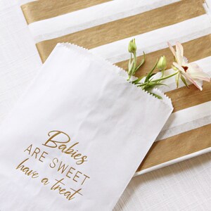 May include: White paper bag with gold lettering that reads "Babies are sweet have a treat". The bag is partially filled with a small bouquet of white flowers.