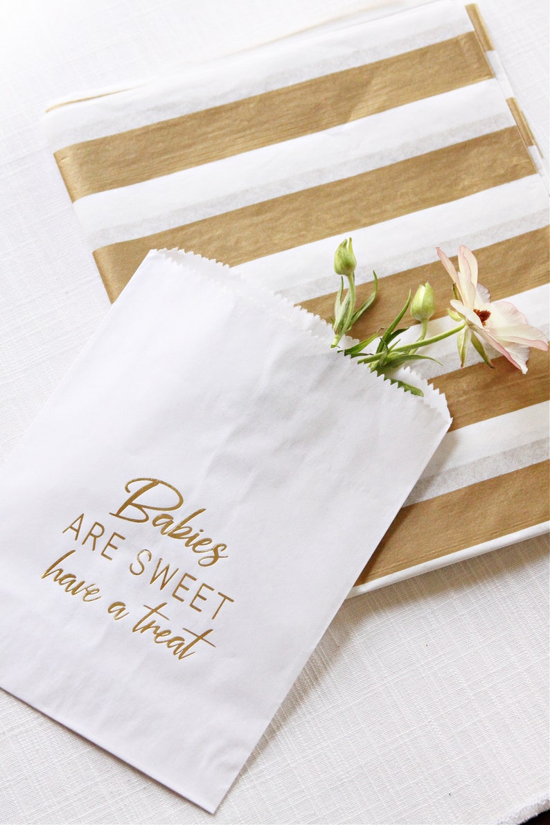 May include: White paper bag with gold lettering that reads "Babies are sweet have a treat". The bag is partially filled with a small bouquet of white flowers.