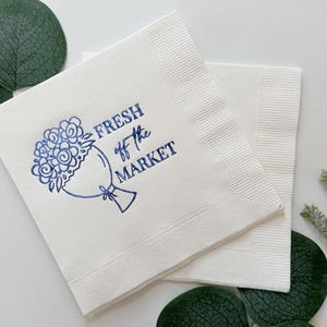 May include: White paper napkins with a blue floral bouquet design and the text "FRESH off the MARKET". The napkins are square and have a textured border. Green leaves and sprigs are in the background.