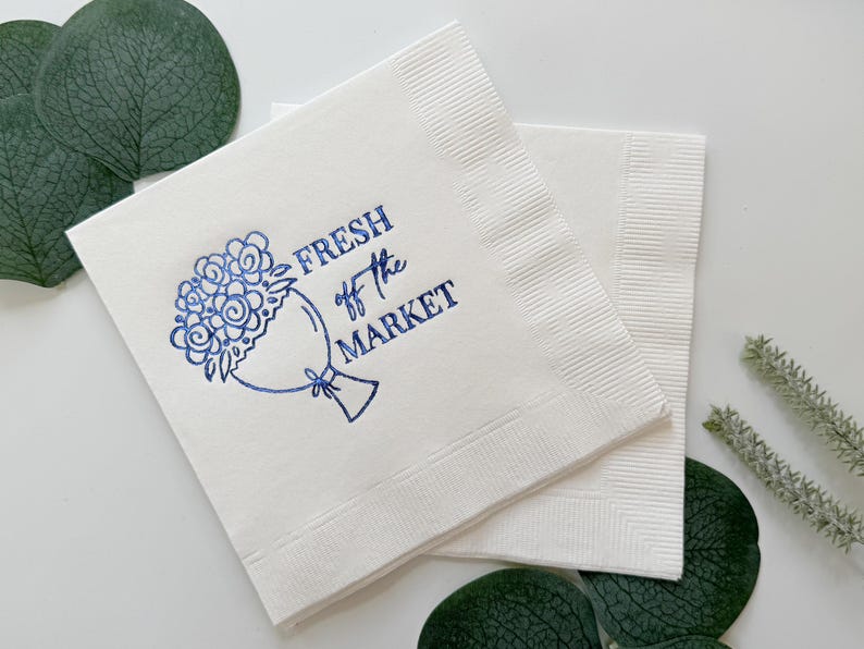 May include: White paper napkins with a blue floral bouquet design and the text "FRESH off the MARKET". The napkins are square and have a textured border. Green leaves and sprigs are in the background.