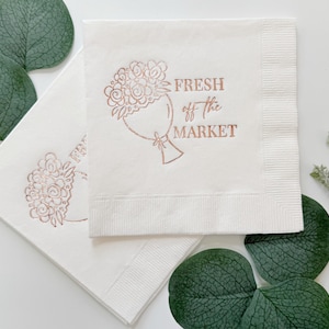 May include: Two white paper napkins with a rose gold floral bouquet design and the text "FRESH off the MARKET". The napkins are surrounded by green leaves and sprigs, creating a fresh and natural aesthetic.