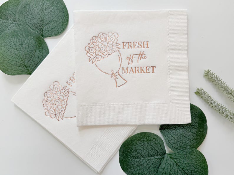 May include: Two white paper napkins with a rose gold floral bouquet design and the text "FRESH off the MARKET". The napkins are surrounded by green leaves and sprigs, creating a fresh and natural aesthetic.