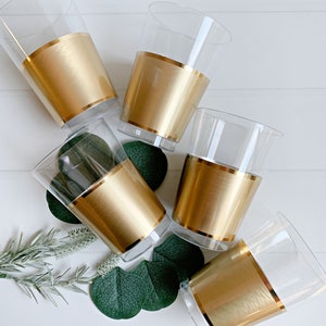 May include: Five clear plastic cups with a gold band around the middle. The cups are arranged on a white surface with green leaves and sprigs of greenery. The gold band adds a touch of elegance to the cups.
