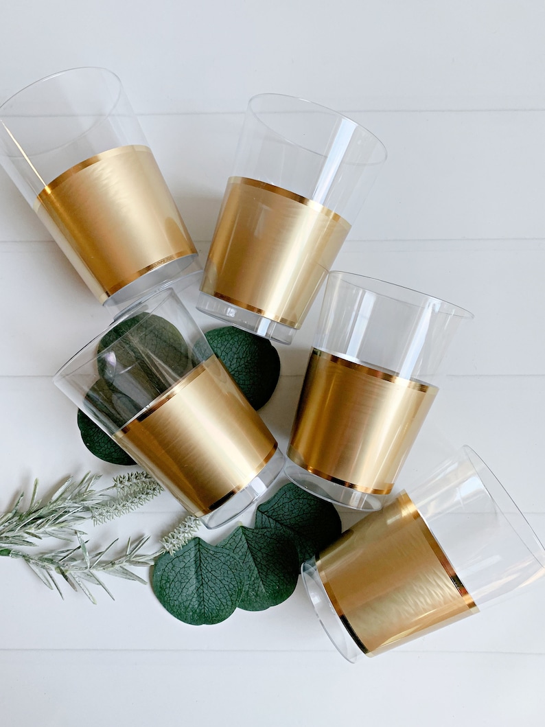 May include: Five clear plastic cups with a gold band around the middle. The cups are arranged on a white surface with green leaves and sprigs of greenery. The gold band adds a touch of elegance to the cups.