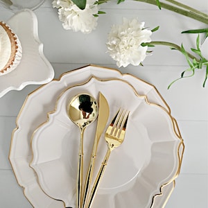 May include: A set of two stacked white scalloped plates with gold-rimmed edges, accompanied by a gold-toned spoon, knife, and fork. The cutlery is arranged neatly in the center of the plates, reflecting light on their polished surfaces.