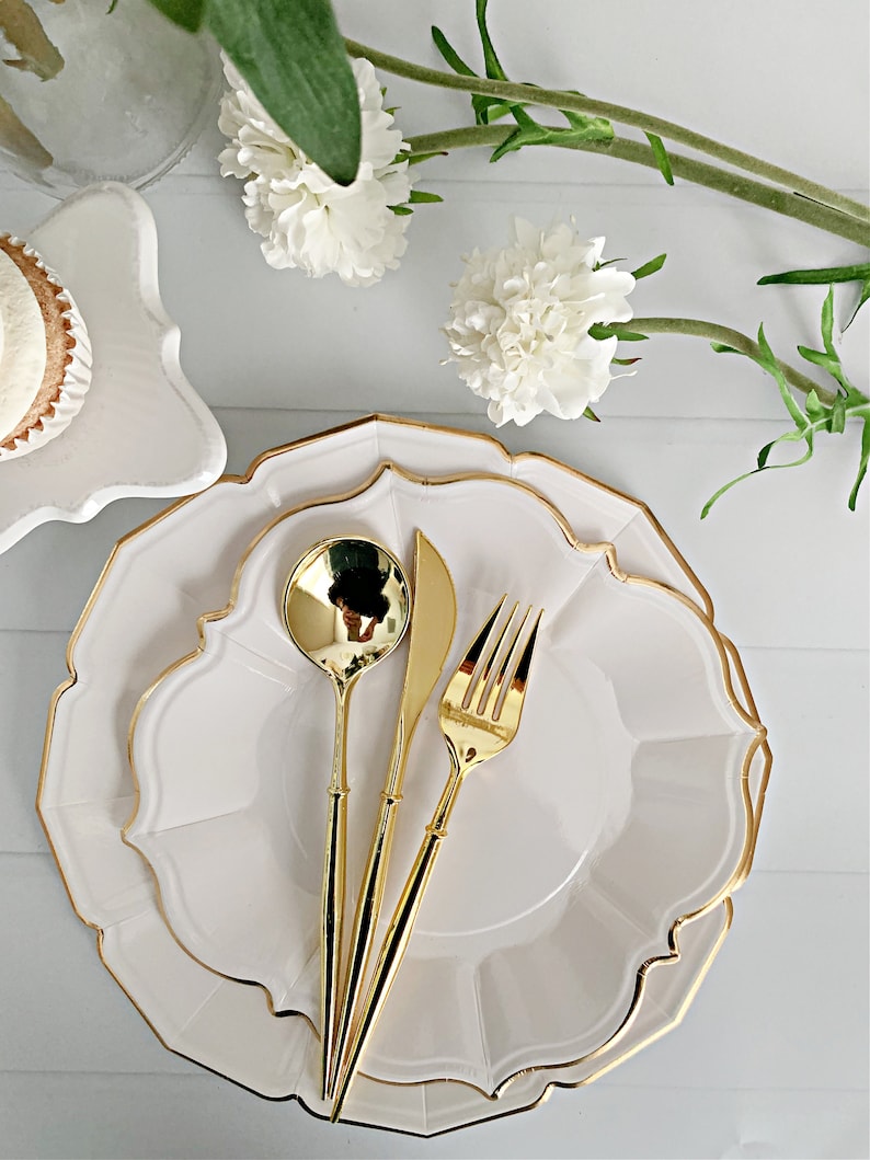 May include: A set of two stacked white scalloped plates with gold-rimmed edges, accompanied by a gold-toned spoon, knife, and fork. The cutlery is arranged neatly in the center of the plates, reflecting light on their polished surfaces.