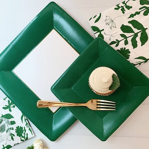 May include: A festive arrangement featuring emerald green square plates and a matching frame. A cupcake with white frosting and a gold fork are on a plate. Napkins with green floral patterns complete the setting, perfect for a party.