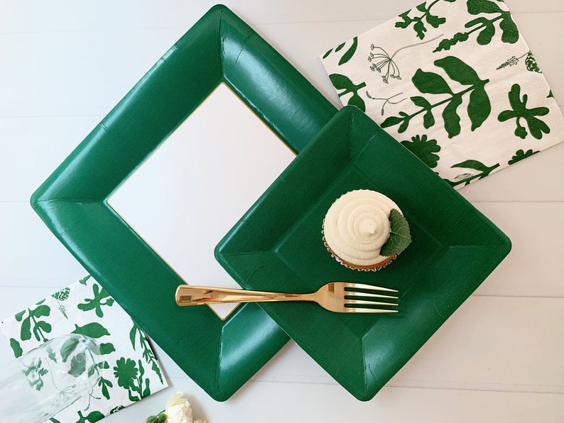May include: A festive arrangement featuring emerald green square plates and a matching frame. A cupcake with white frosting and a gold fork are on a plate. Napkins with green floral patterns complete the setting, perfect for a party.