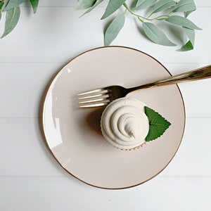 May include: A cupcake with white frosting and a green leaf on a pink plate with a gold rim. A gold fork rests on the plate. The scene is set against a white background with green foliage.