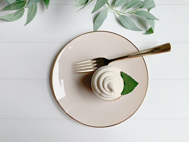 May include: A cupcake with white frosting and a green leaf on a pink plate with a gold rim. A gold fork rests on the plate. The scene is set against a white background with green foliage.