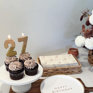 May include: Four chocolate cupcakes with light brown frosting on a white pedestal stand, topped with two lit gold glitter candles forming the number 27. Next to the stand is a white plate with the text HAPPY BIRTHDAY in gold lettering.