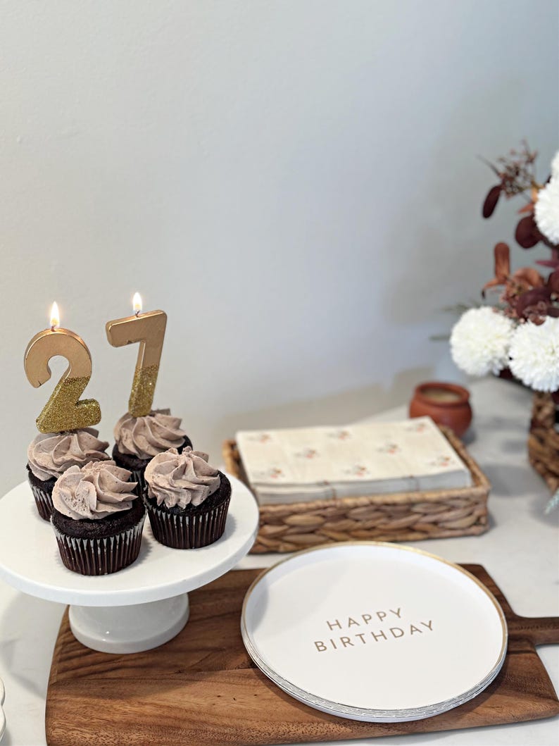 May include: Four chocolate cupcakes with light brown frosting on a white pedestal stand, topped with two lit gold glitter candles forming the number 27. Next to the stand is a white plate with the text HAPPY BIRTHDAY in gold lettering.