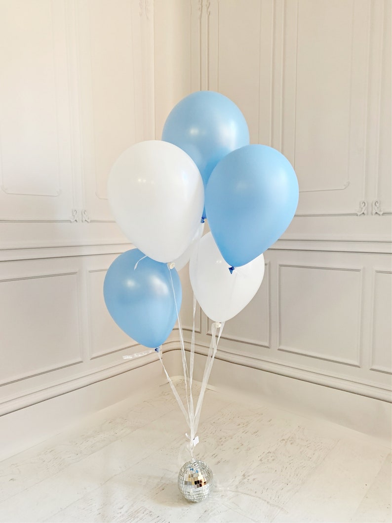 May include: A cluster of five round balloons in alternating shades of light blue and white, tied together with white ribbons. The balloons are anchored at the base by a small, spherical, silver mirrored disco ball with a reflective surface.