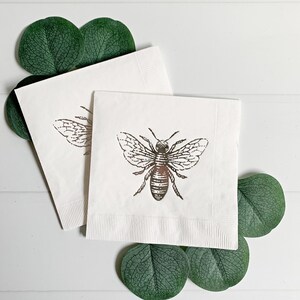 May include: Two white paper napkins with a gold bee design. The napkins are folded and surrounded by green eucalyptus leaves.