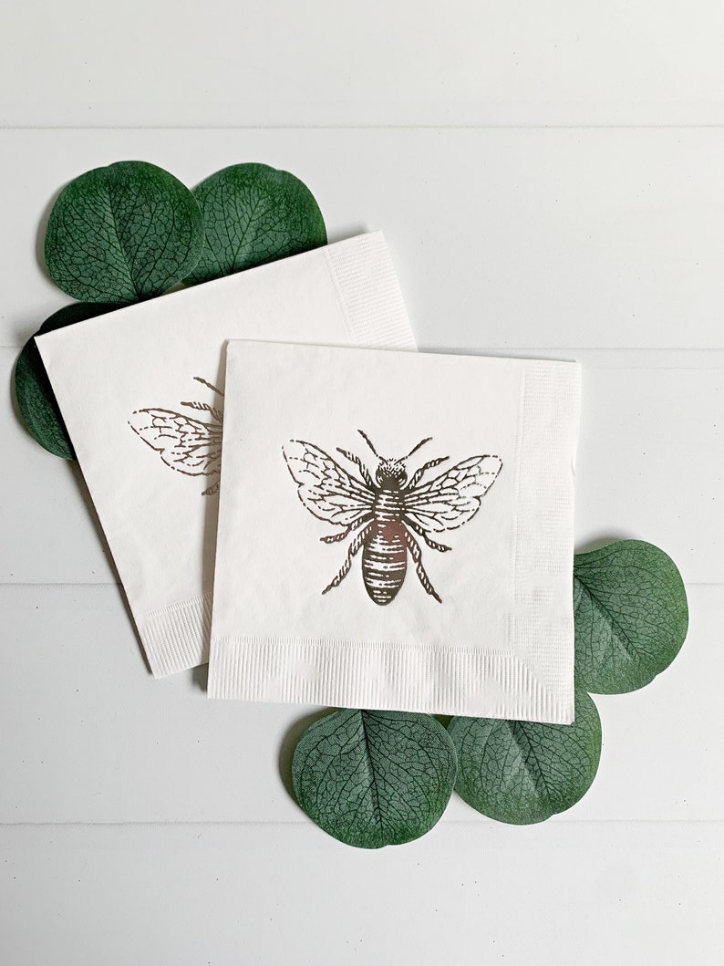 May include: Two white paper napkins with a gold bee design. The napkins are folded and surrounded by green eucalyptus leaves.