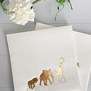 May include: White paper napkins with gold foil animal silhouettes. The animals include a lion, elephant, and giraffe.