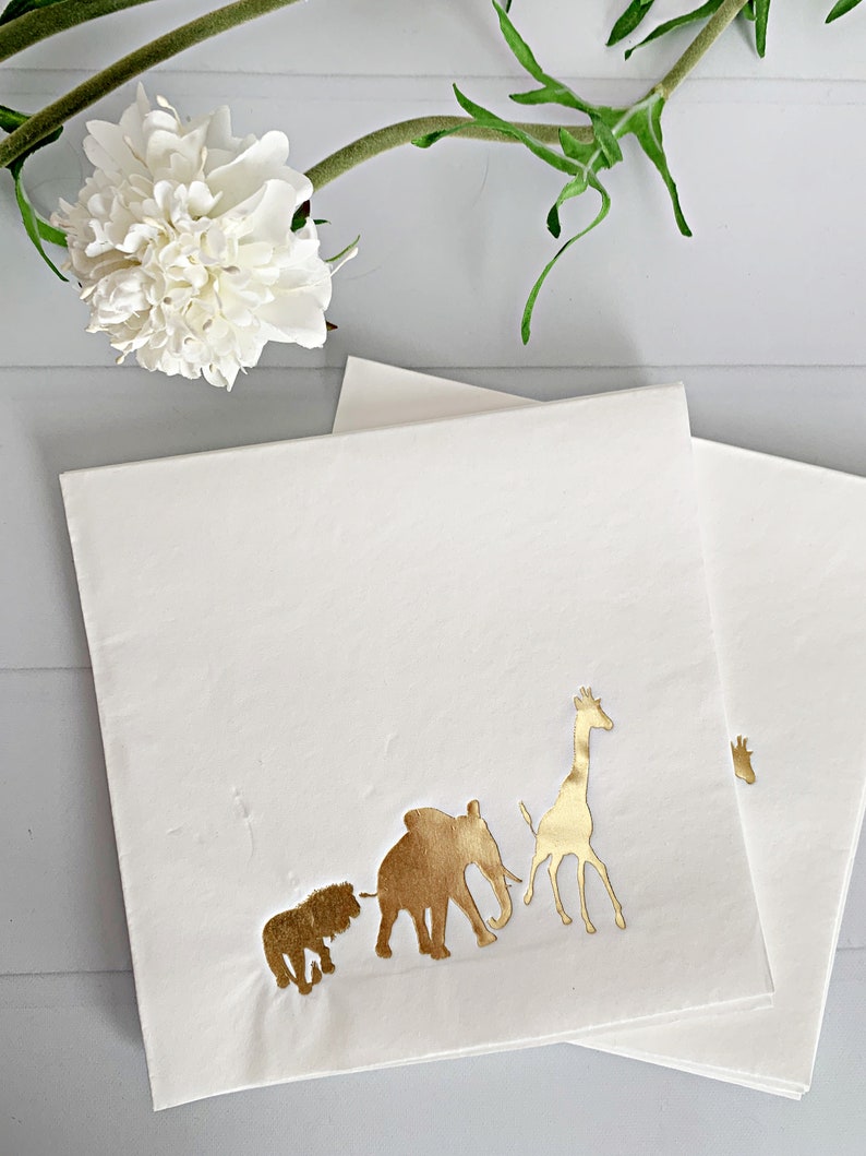 May include: White paper napkins with gold foil animal silhouettes. The animals include a lion, elephant, and giraffe.