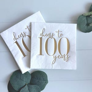 May include: Two white square napkins with gold text that reads "cheers to 100 years." The napkins are arranged on a white surface with green leaves. The napkins are folded and ready for use.